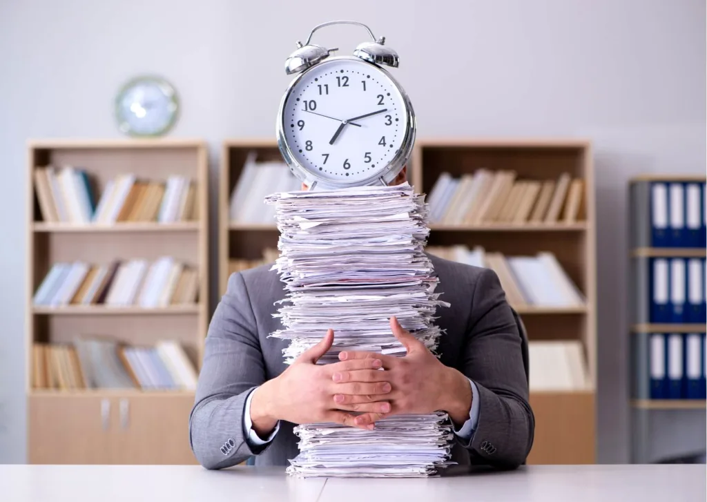Employee behind a tall stack of paperwork with an alarm clock, waiting on a scheduled shift